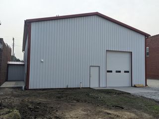Gray metal warehouse with a large garage door and small side door on a gravel lot.
