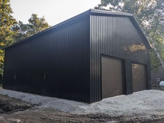 Black metal barn with sloped roof and roll-up doors on a gravel lot, viewed from the side.