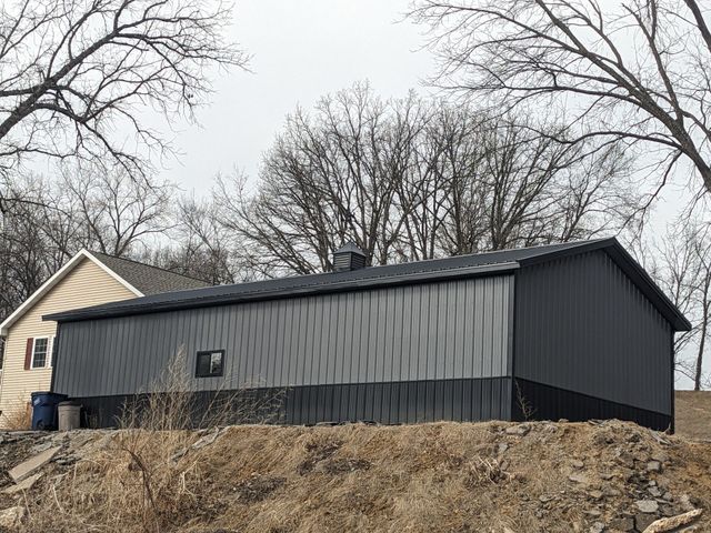 Dark gray shed with slatted siding on a rocky hillside beside a beige house, under bare winter trees