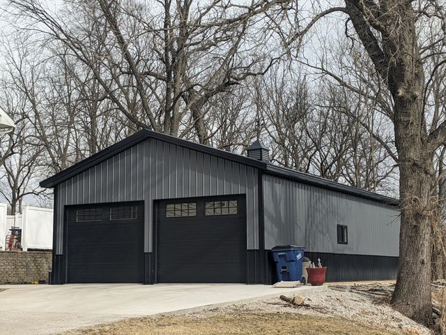 Gray garage with two black doors, blue and red bins, and leafless trees in front.