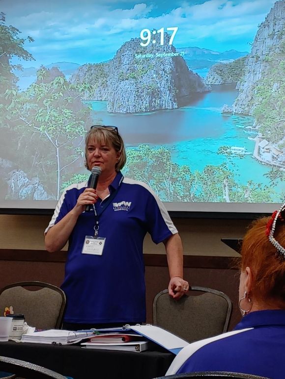 Woman speaking into a microphone at a presentation; a tropical landscape is on the screen behind her.