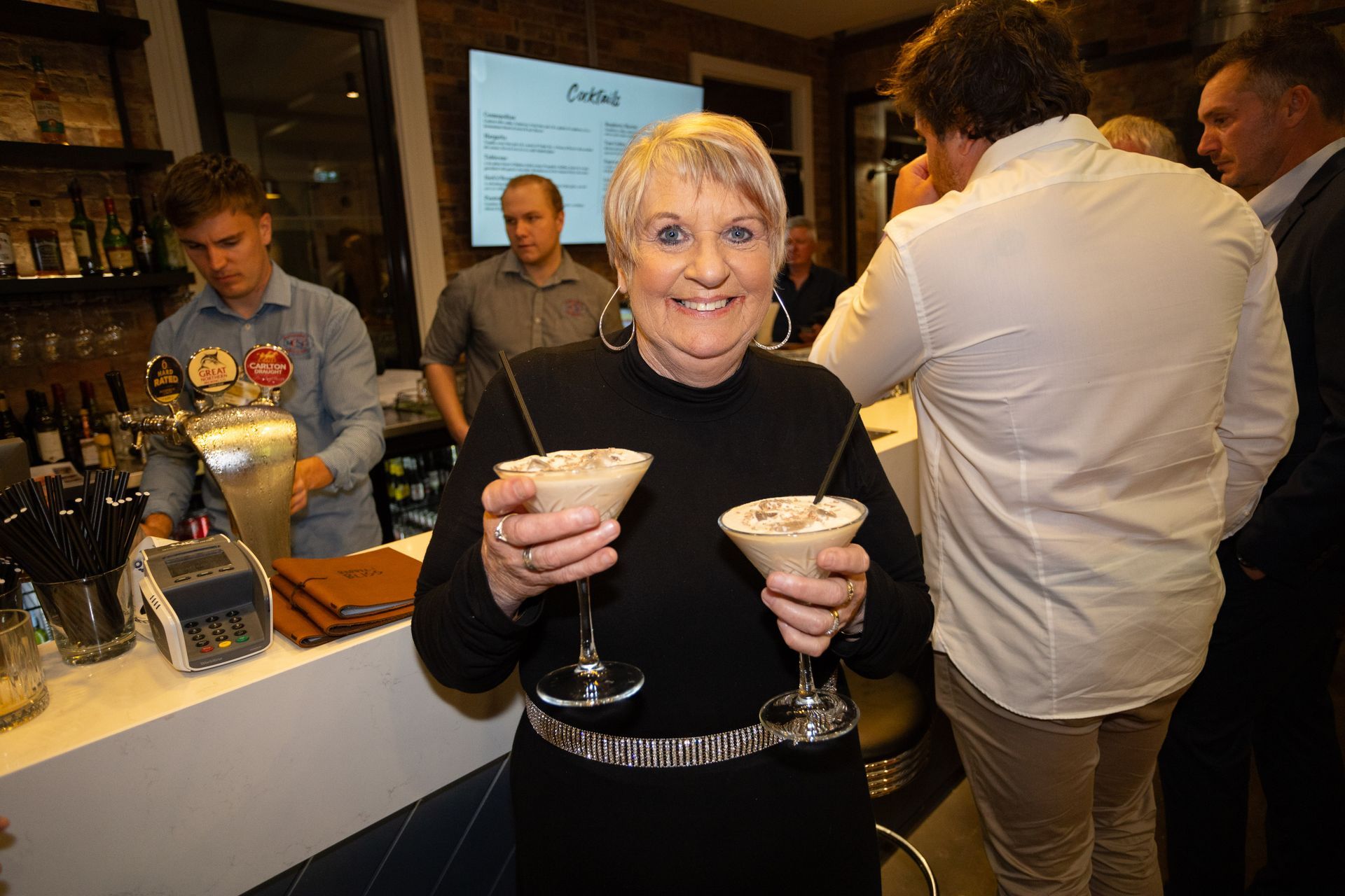 Woman holding two cocktails, smiling, in a bar setting. Others are present.