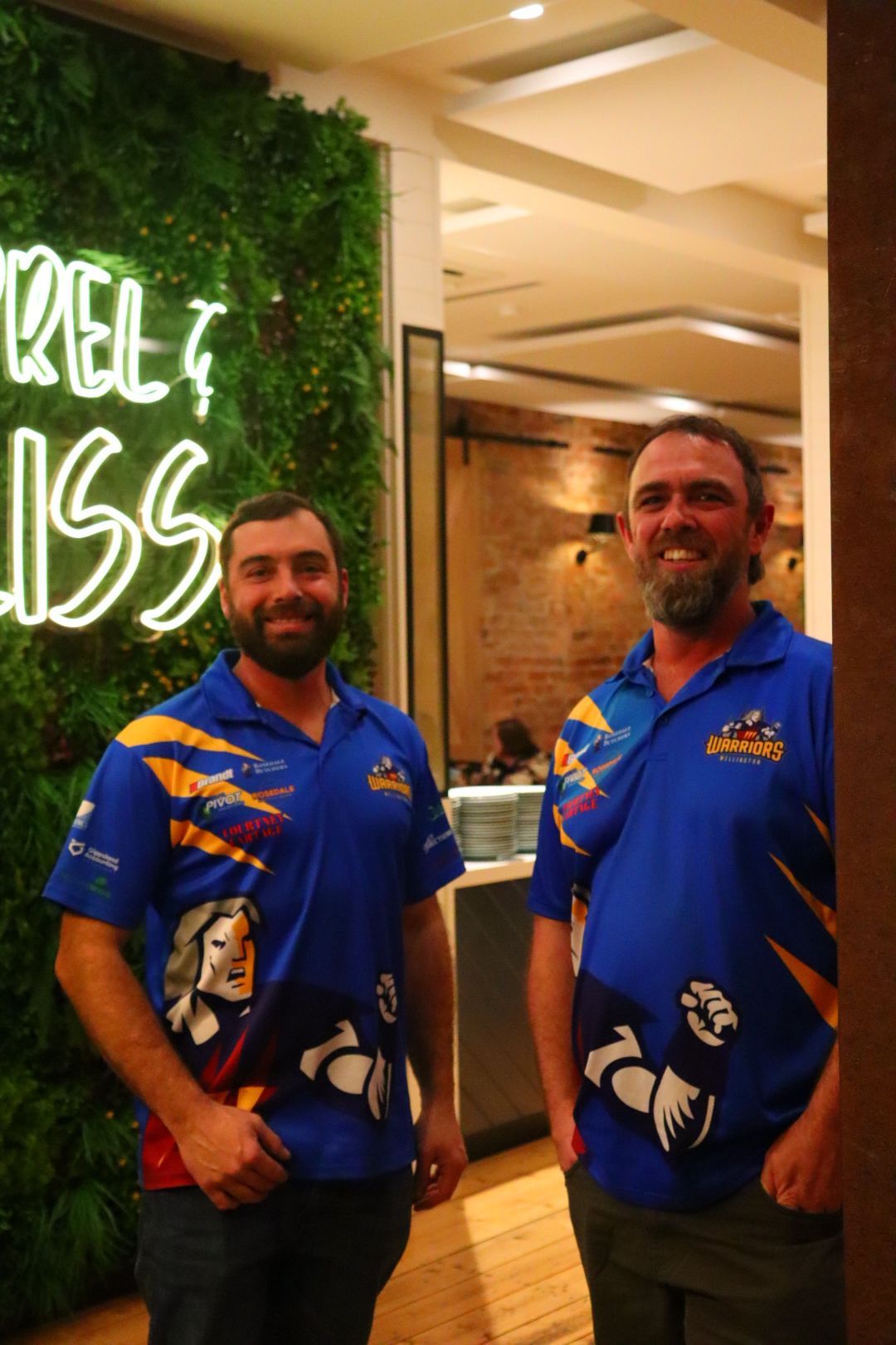 Two men in blue shirts stand in front of a green wall with neon lettering.