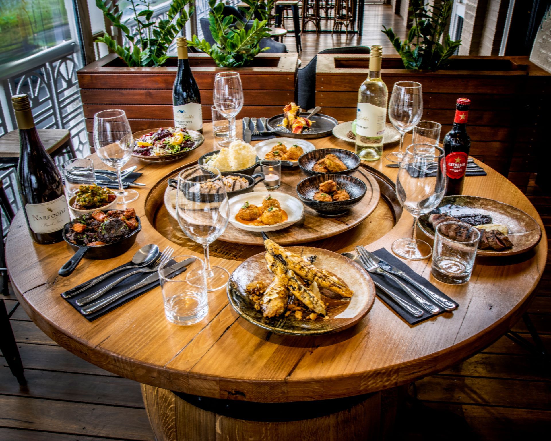 Round wooden table set with food, drinks, and glassware at a restaurant.