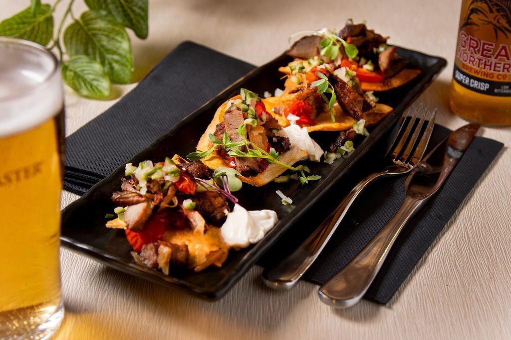 Trio of tostadas topped with meat, vegetables, and sauce on a black plate with silverware, beer, and a napkin.