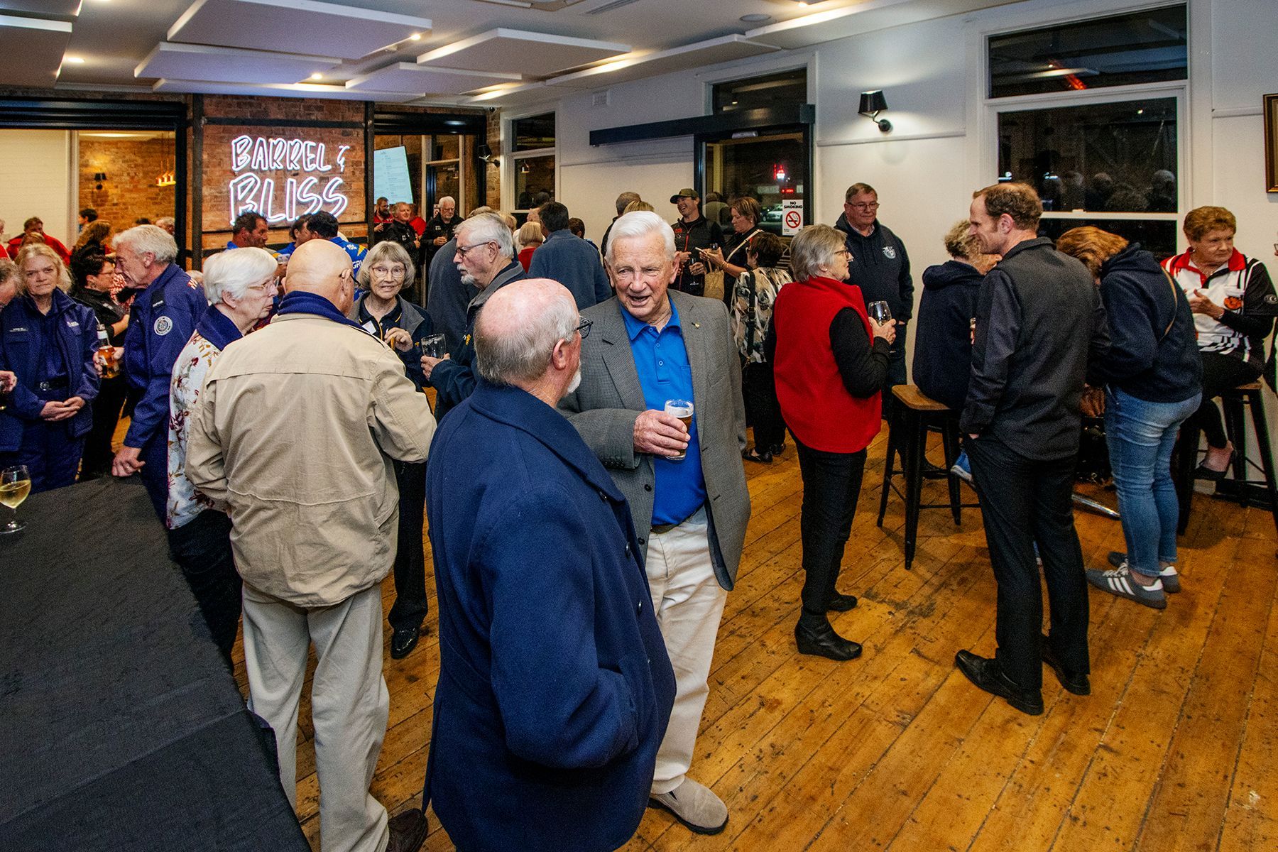 People gathered at an event in a wood-floored room, some drinking and chatting; a sign says 