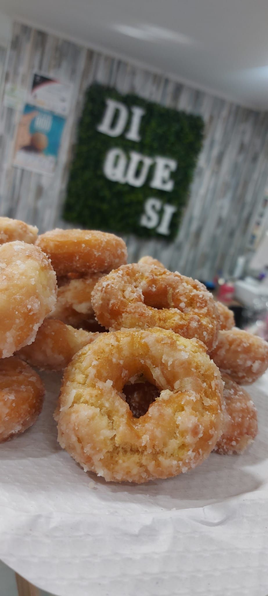 Primer plano de donas cubiertas de azúcar apiladas sobre una superficie blanca, con un cartel verde que dice
