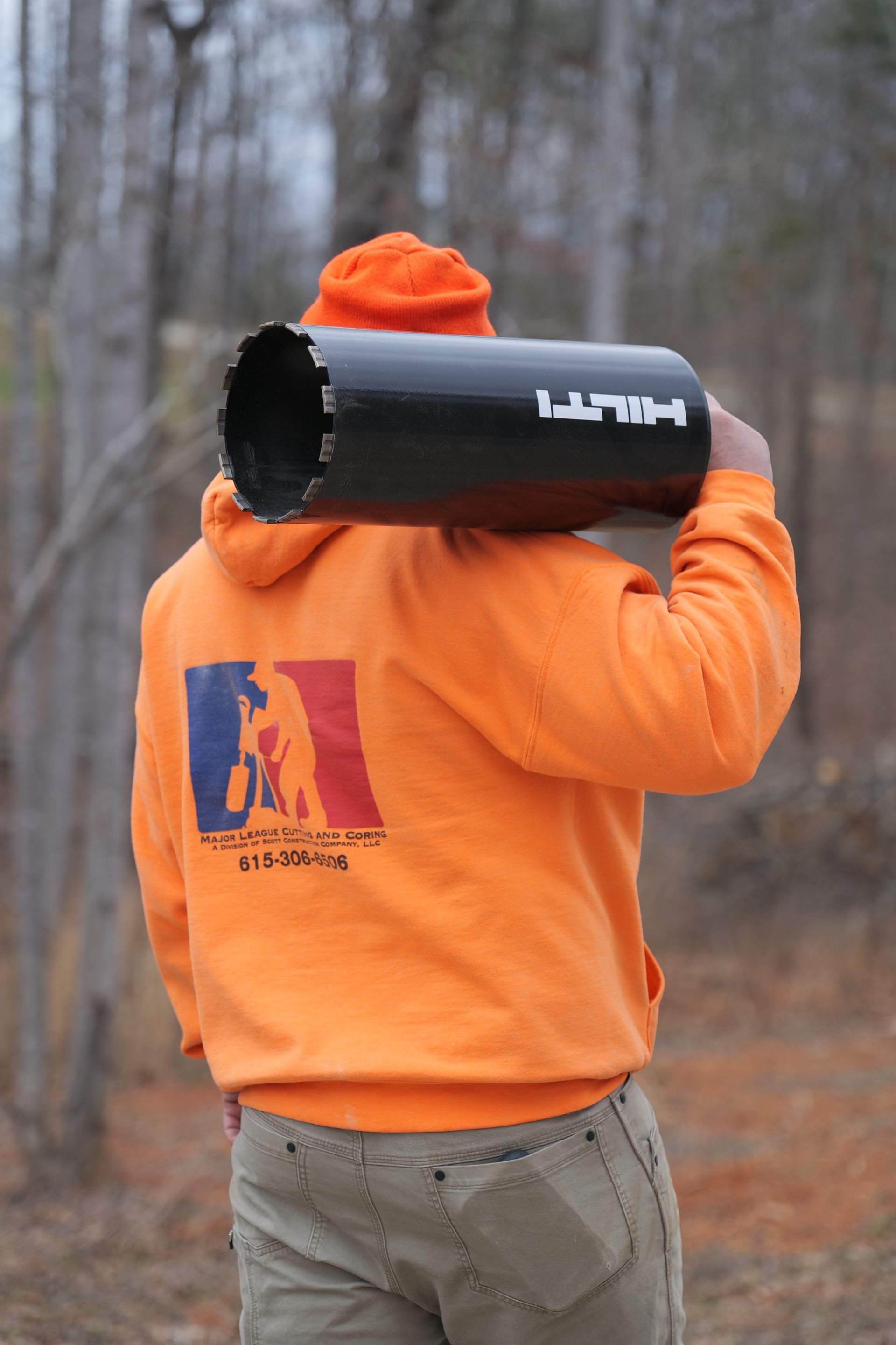 A man in an orange hoodie is carrying a large black drill bit on his shoulder.