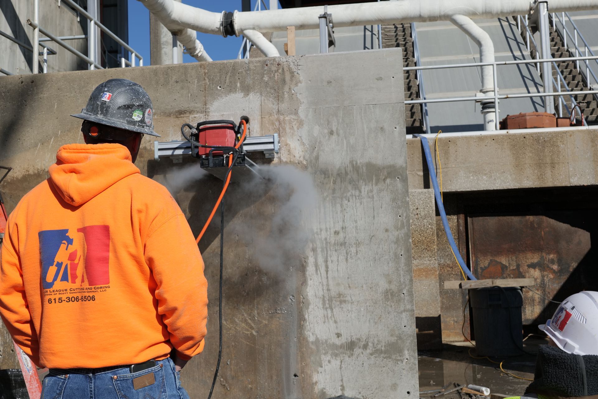 A man wearing a hard hat and an orange hoodie is cutting a concrete wall