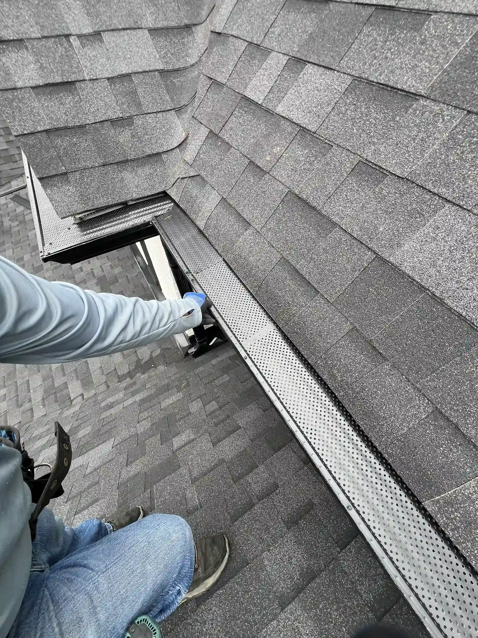 Person on roof installing gutter guard. Gray shingles, silver gutter guard, cutting tool.