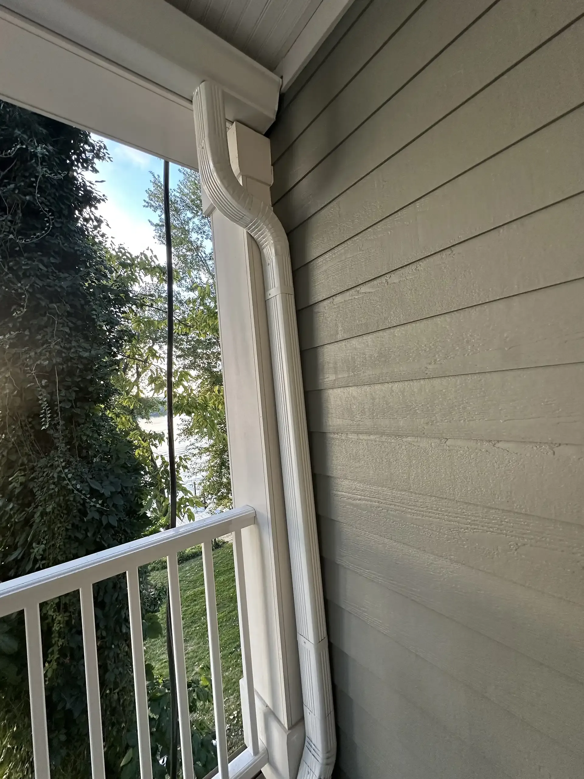 White gutter along a light gray siding on a balcony, with a white railing. Trees and a body of water in the background.