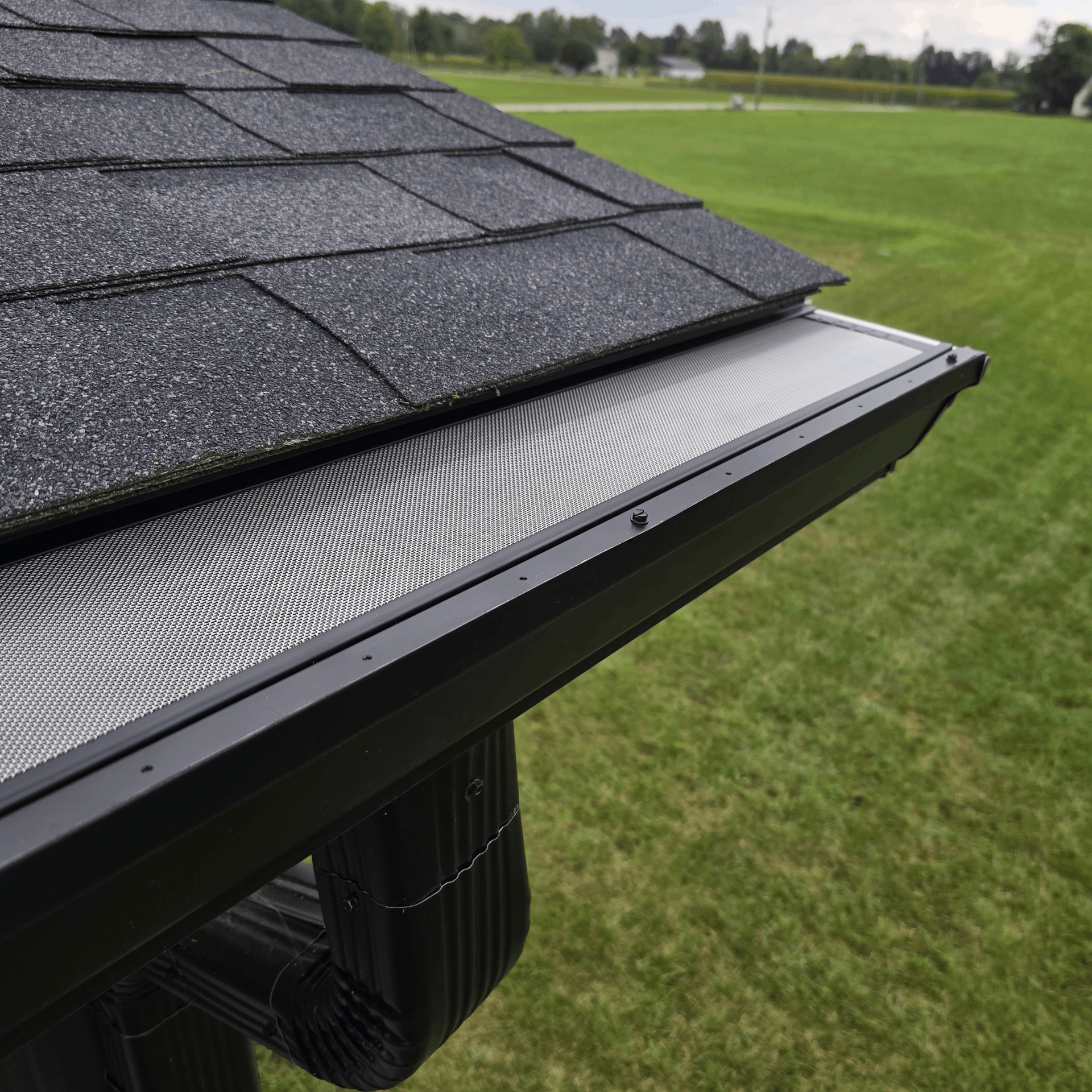 Close-up of a roof edge with gutter guard, black shingles, and downspout over a green lawn