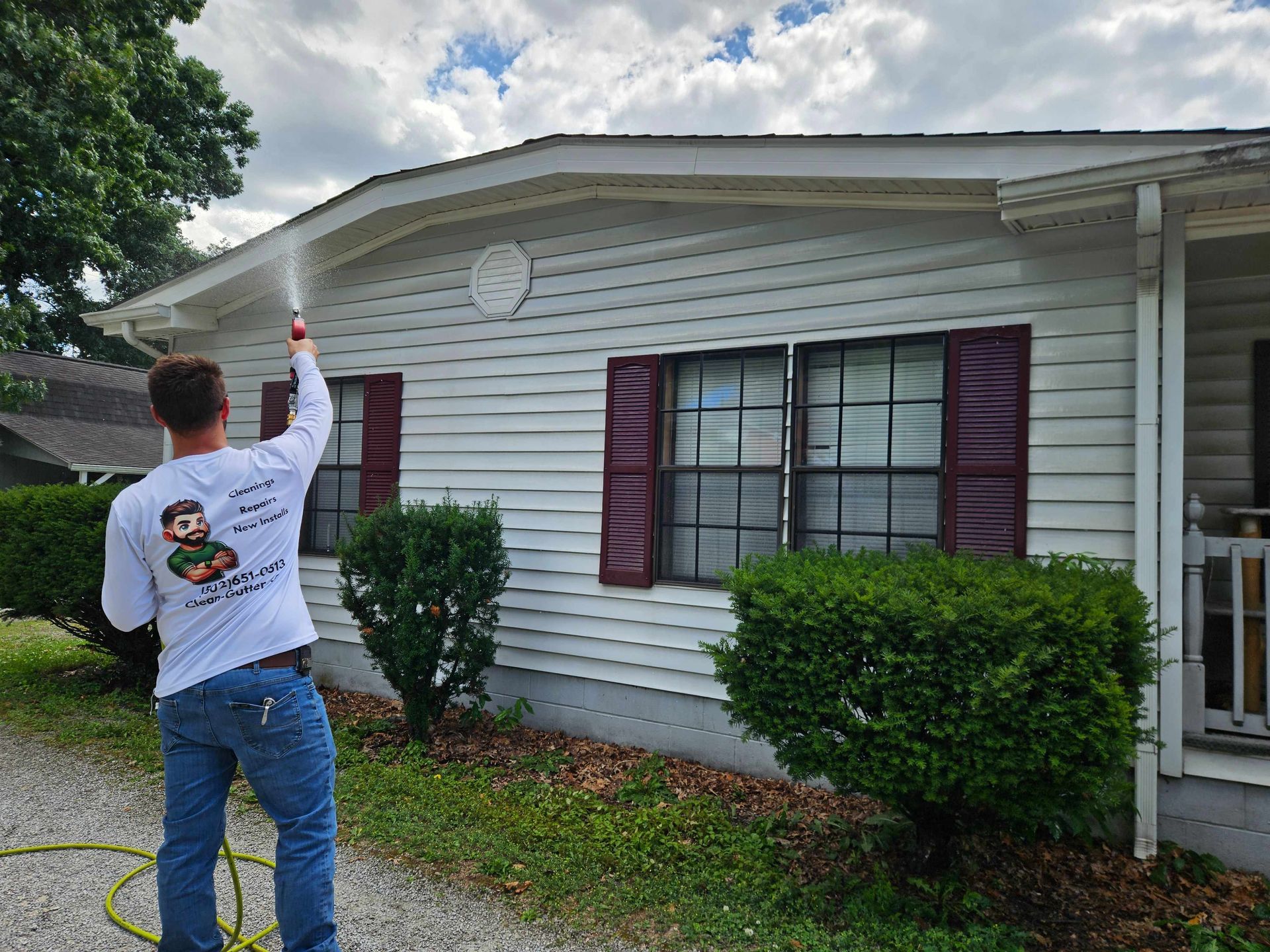 Person on a ladder painting the white exterior siding of a house with maroon shutters.
