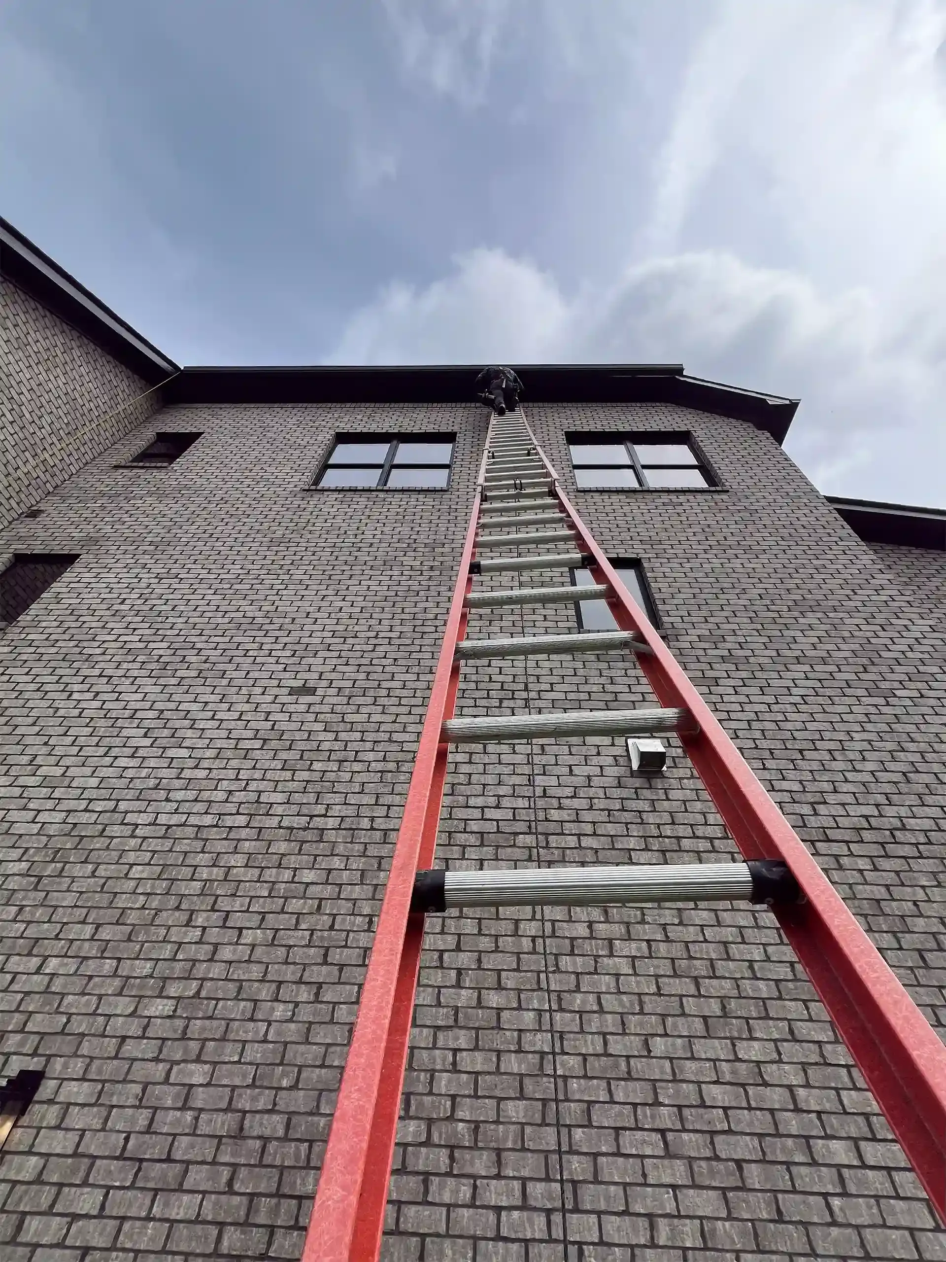 Red ladder against a brick building reaching the roof under a cloudy sky.