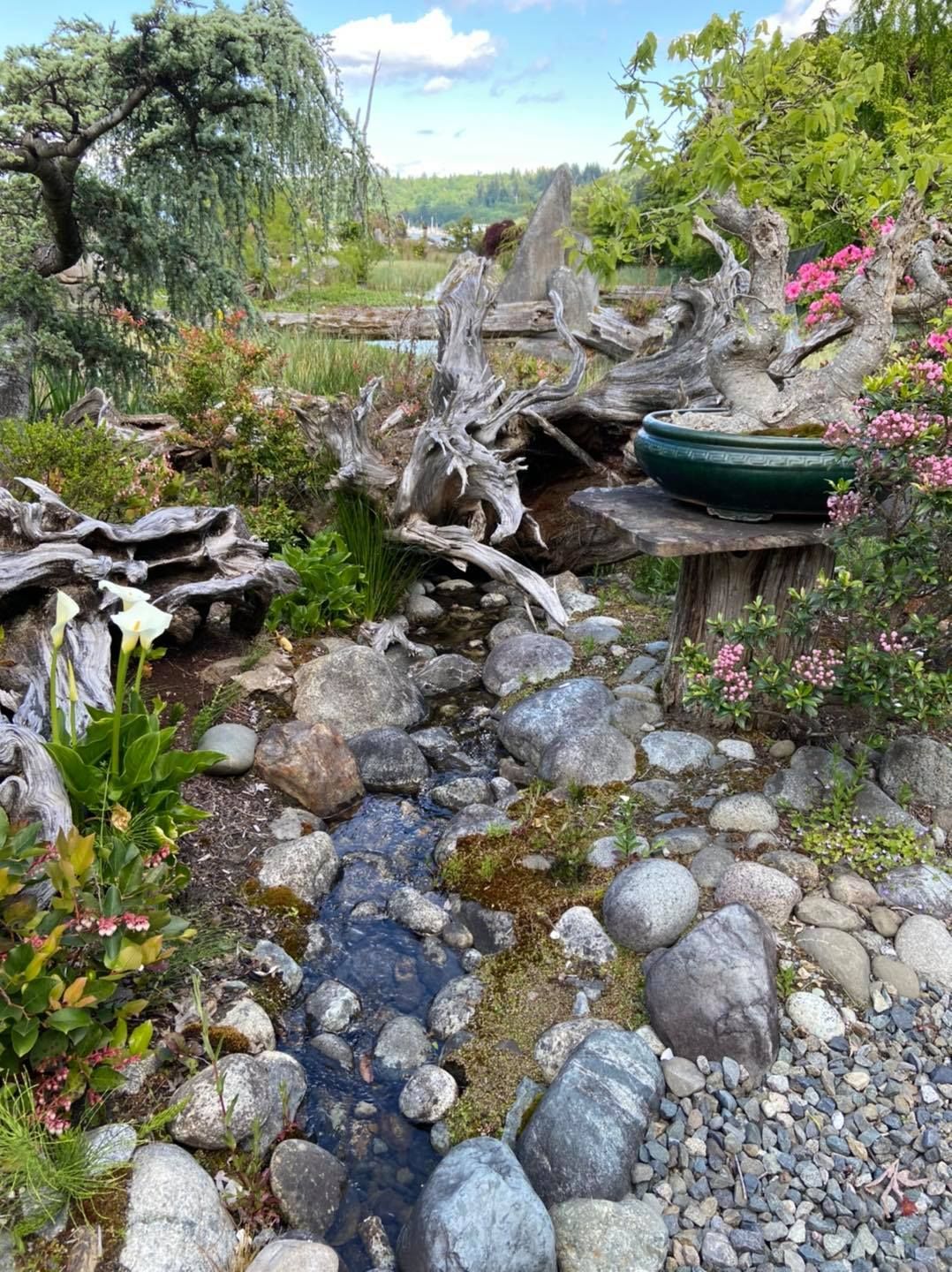 Rock garden path with driftwood, stones, and a green planter amid flowers and greenery