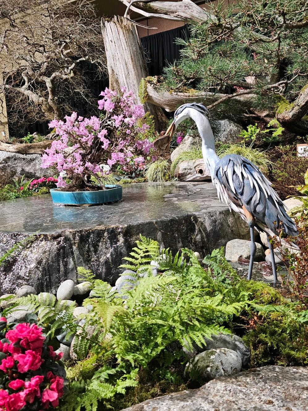 Heron statue beside a garden waterfall with pink flowers and lush ferns