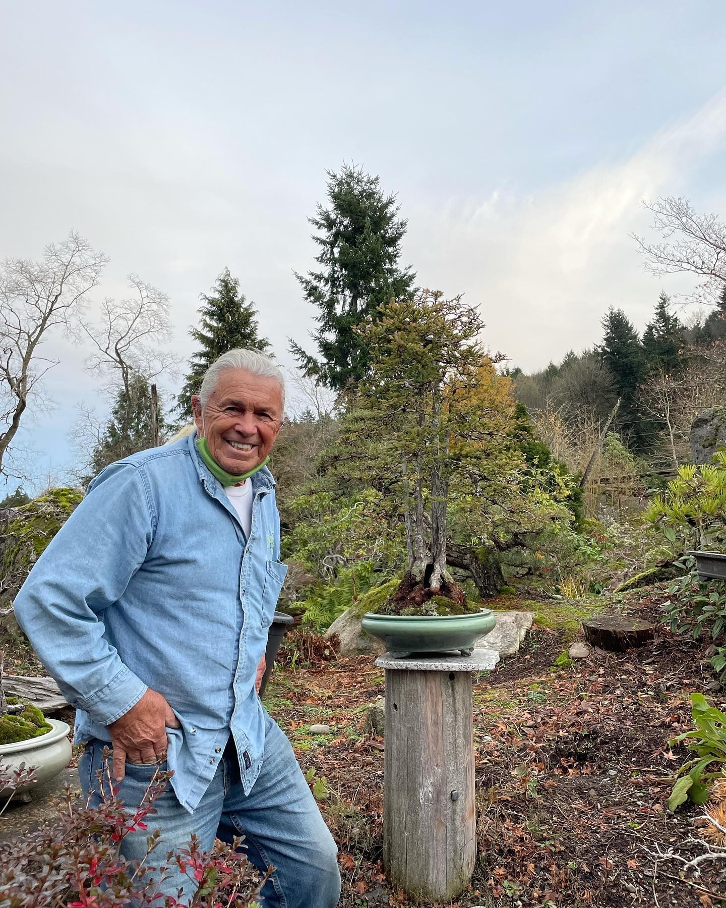 Smiling man in a light blue outfit standing in a garden with potted plants and trees