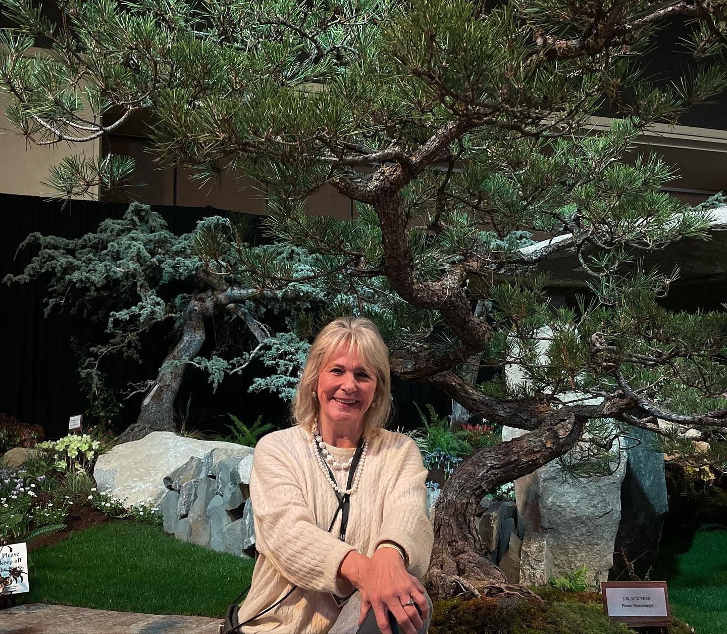 Woman smiling in front of a bonsai tree display and rocks in a garden setting