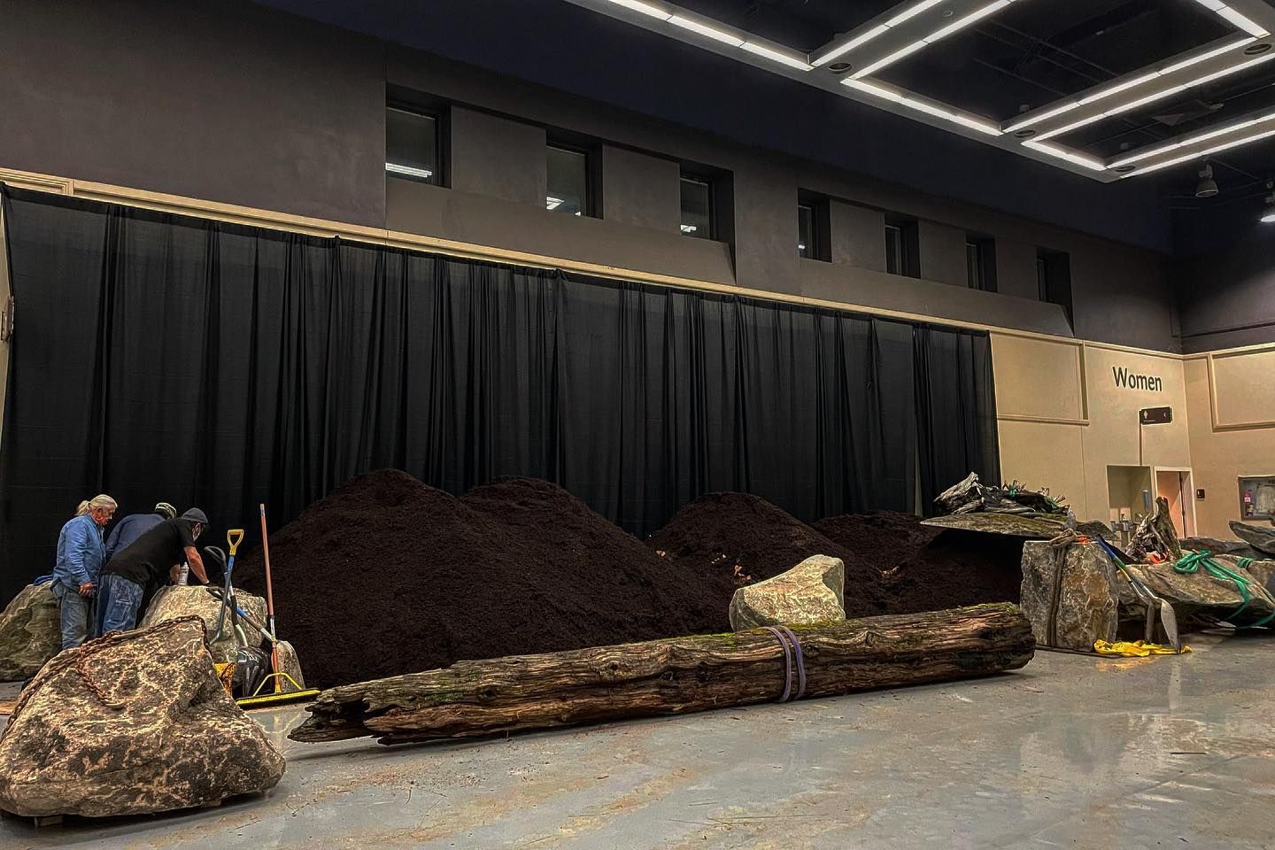 Indoor stage with black drapes, large piles of mulch or debris, and people working at left and right.