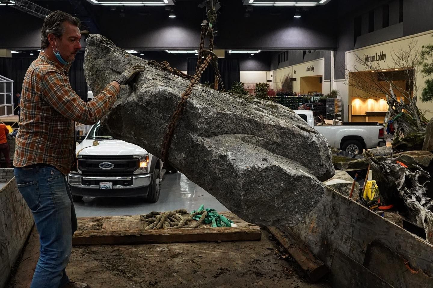 Man in plaid shirt lifting a large gray boulder at an indoor rock display, with a car and buildings behind him