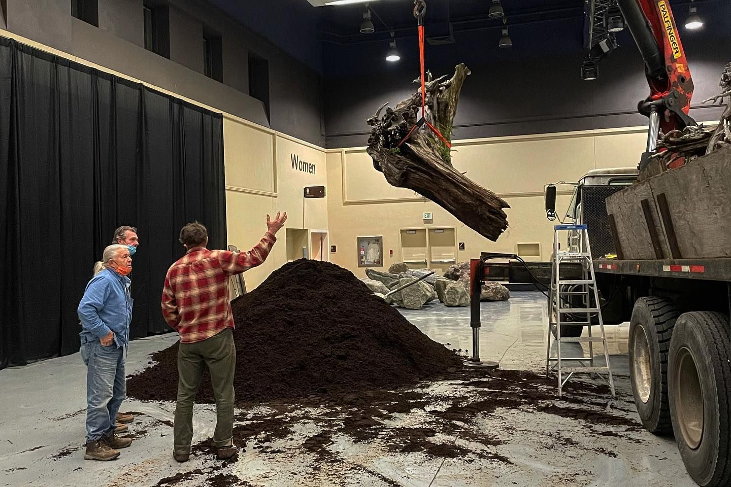 Workers unload a large pile of dark mulch from a truck inside a warehouse.