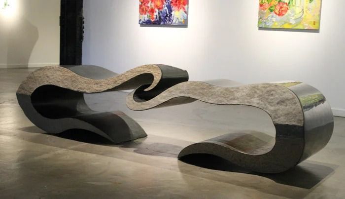 Abstract stone-and-metal bench sculpture in a gallery with colorful paintings on the wall.