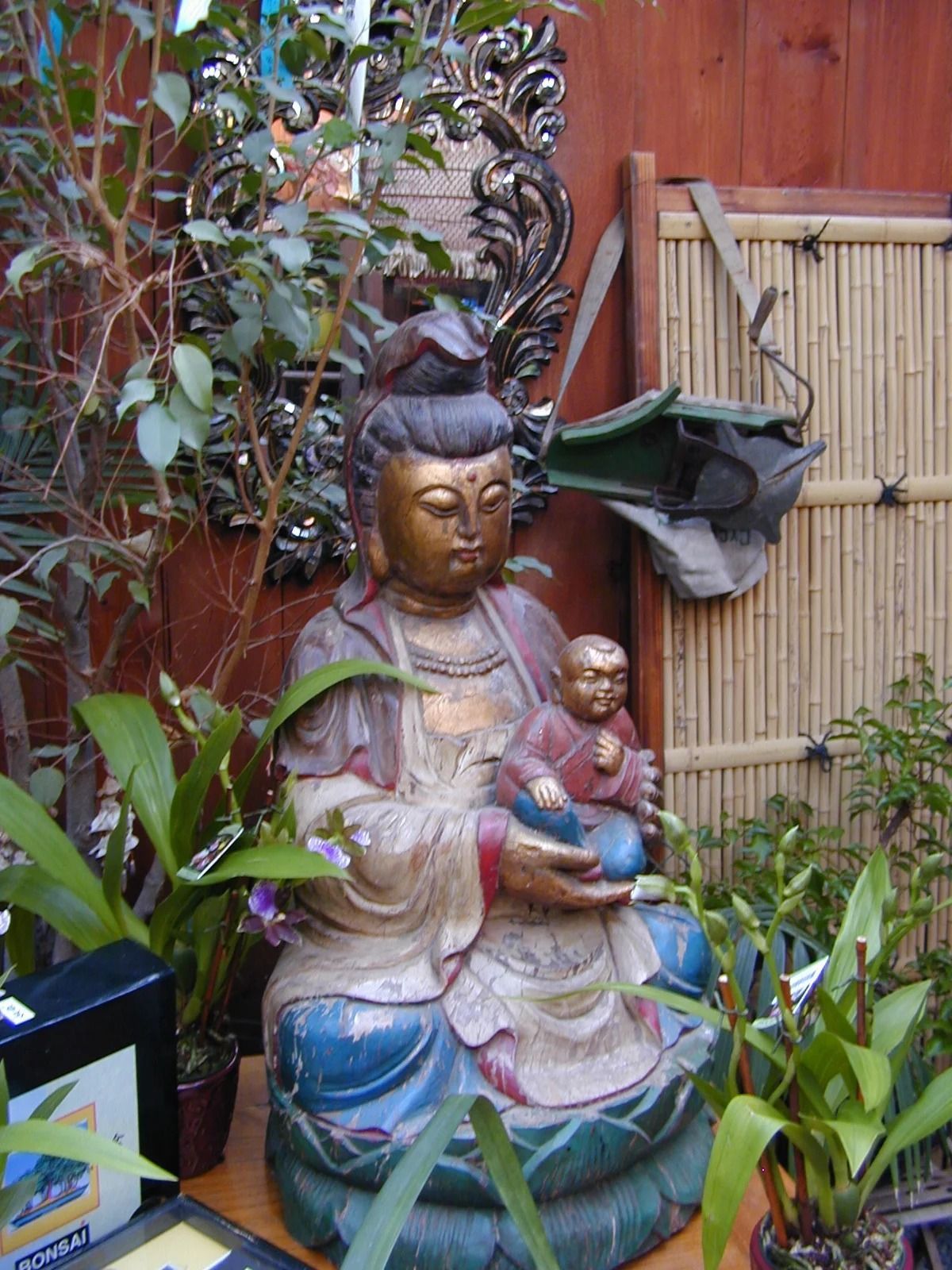 Buddha statue with a small child figure, surrounded by potted plants in a garden setting