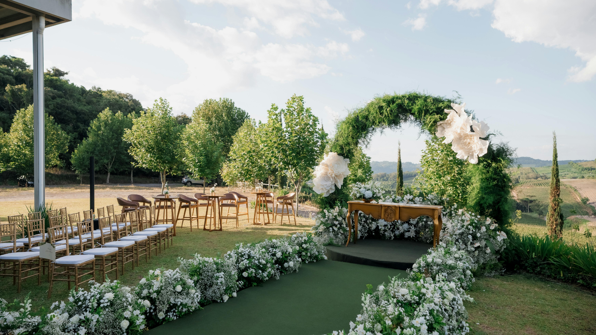 Outdoor wedding ceremony setup with floral arch, white chairs, and greenery under a clear sky
