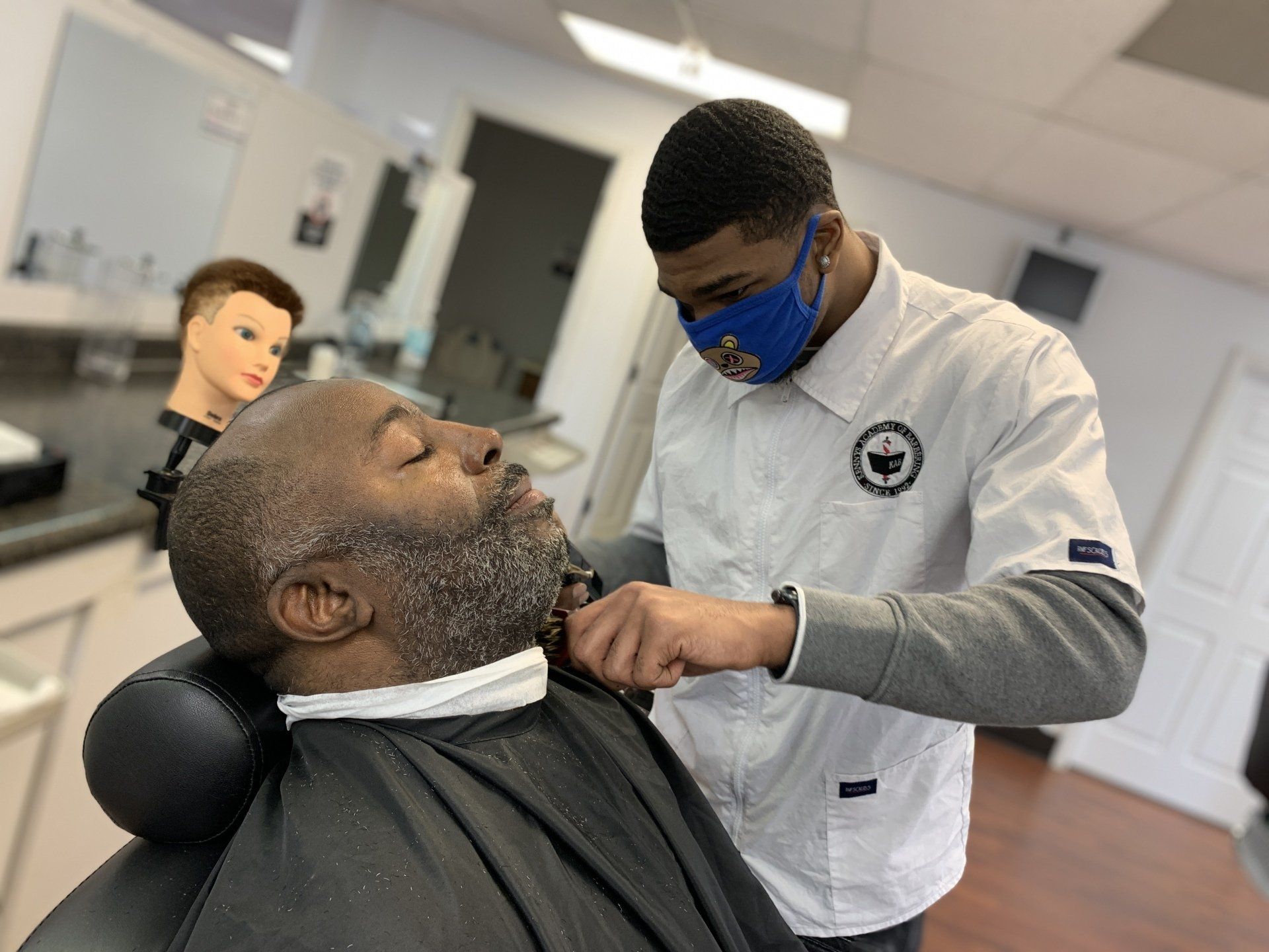 A man is getting his beard shaved by a barber wearing a mask.