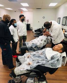 A group of men are standing around a man getting his hair cut in a barber shop.