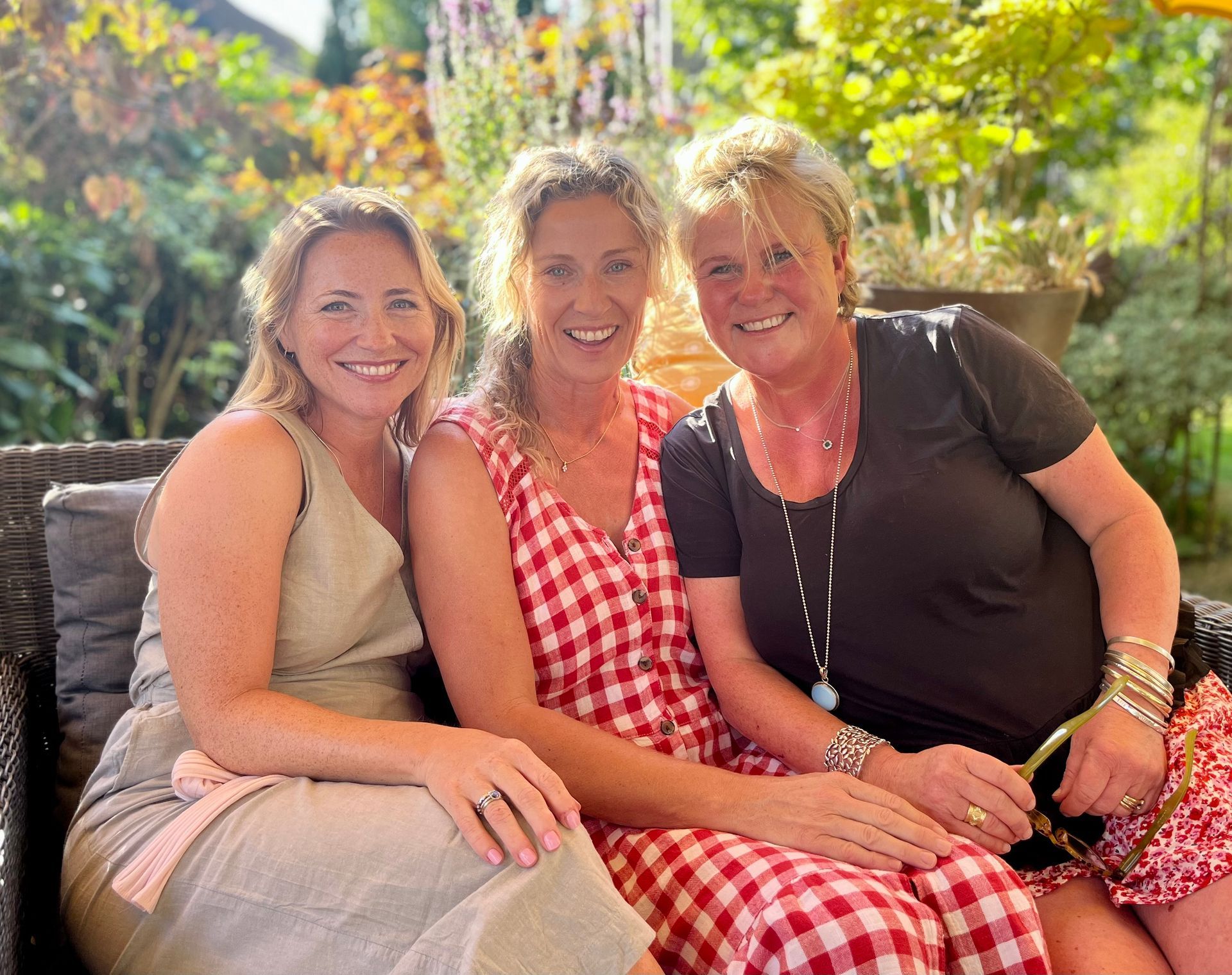 Three women sitting closely together on a couch, smiling outdoors in warm sunlight.