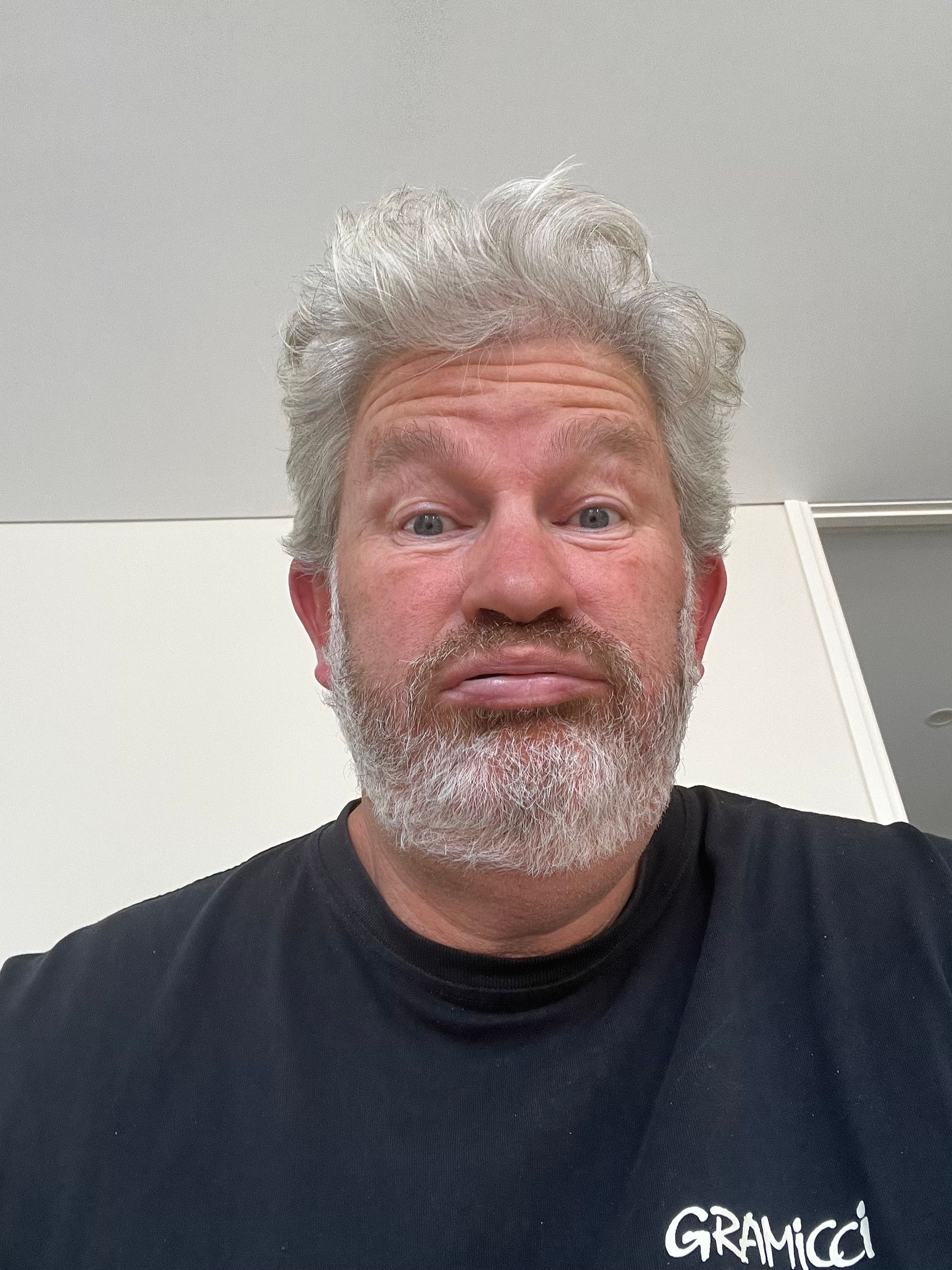 Selfie of a man with light curly hair and a white beard, making a playful face indoors.