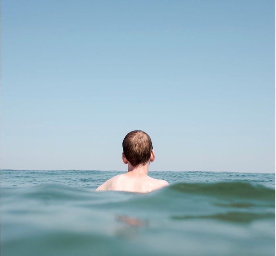 Boy  swimming in ocean, facing away, head above water, blue sky.