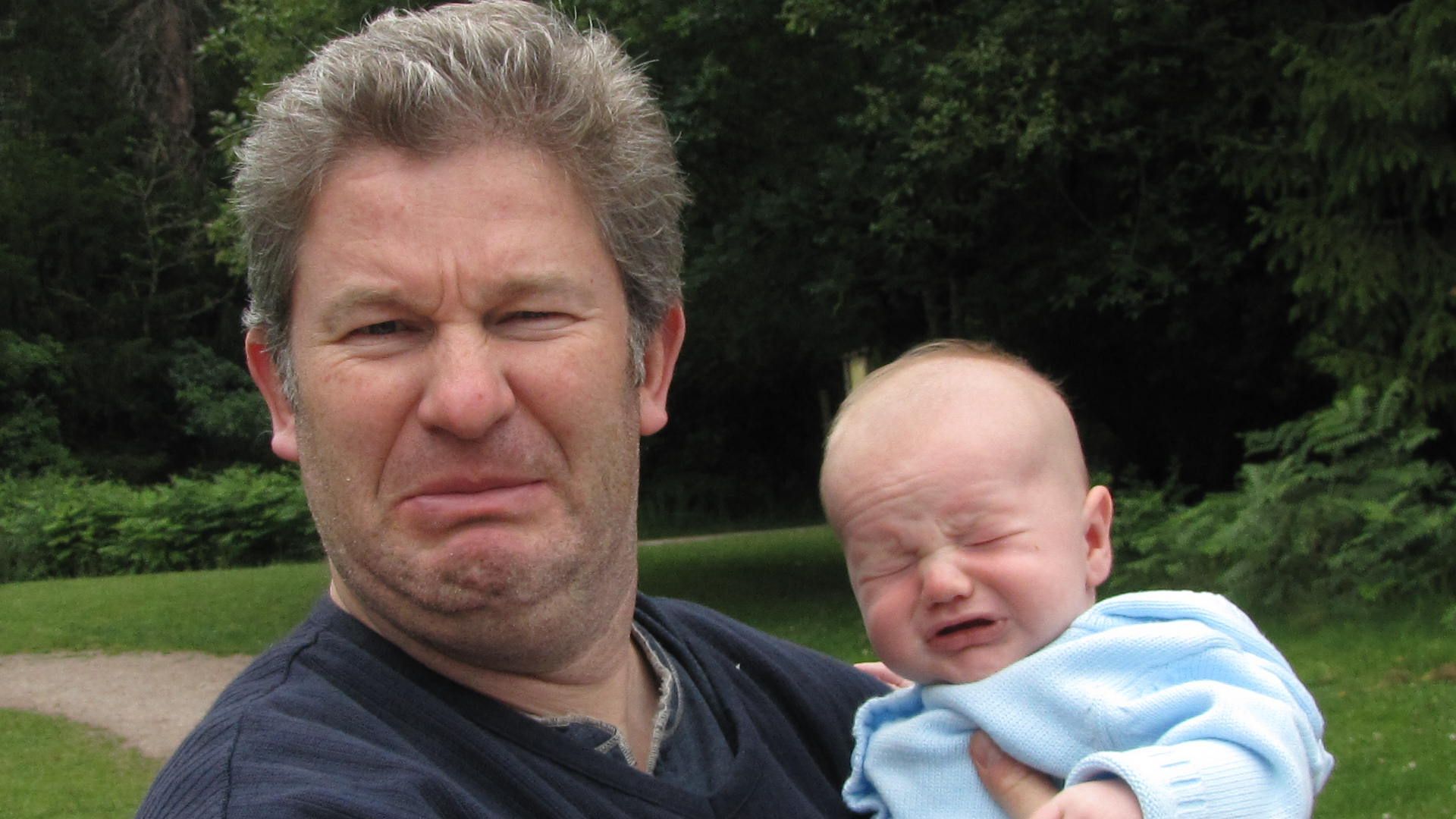 Man holding a crying baby outdoors with trees in the background