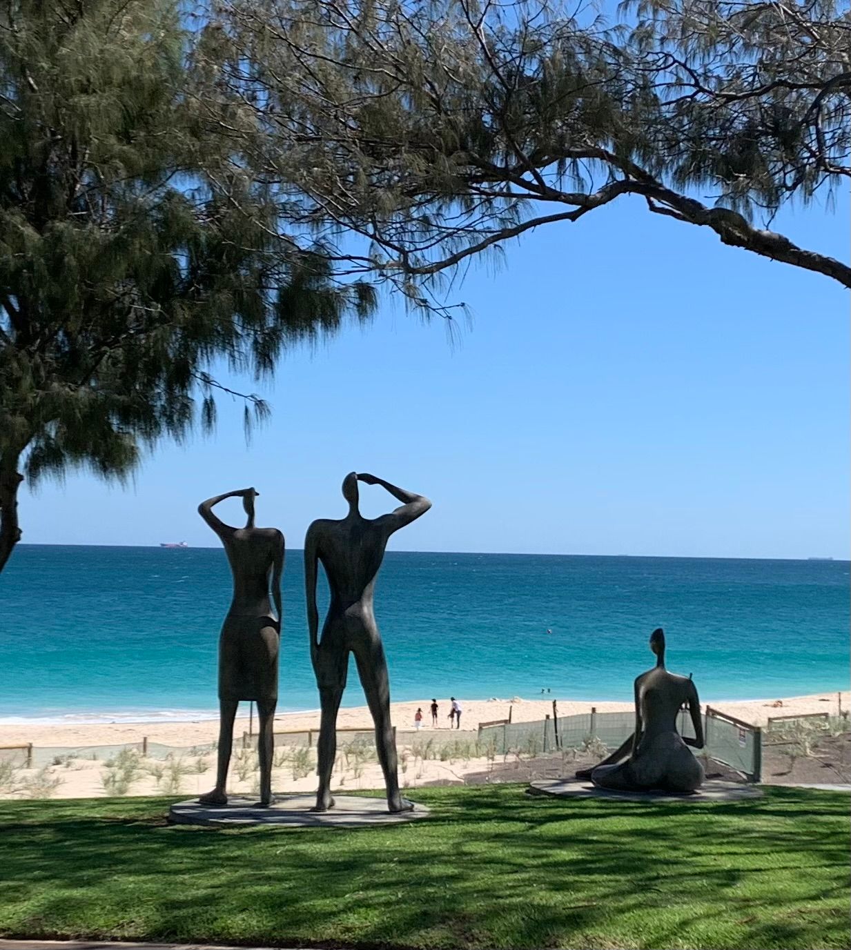 Bronze statues saluting on a grassy beach overlook turquoise water under a blue sky.