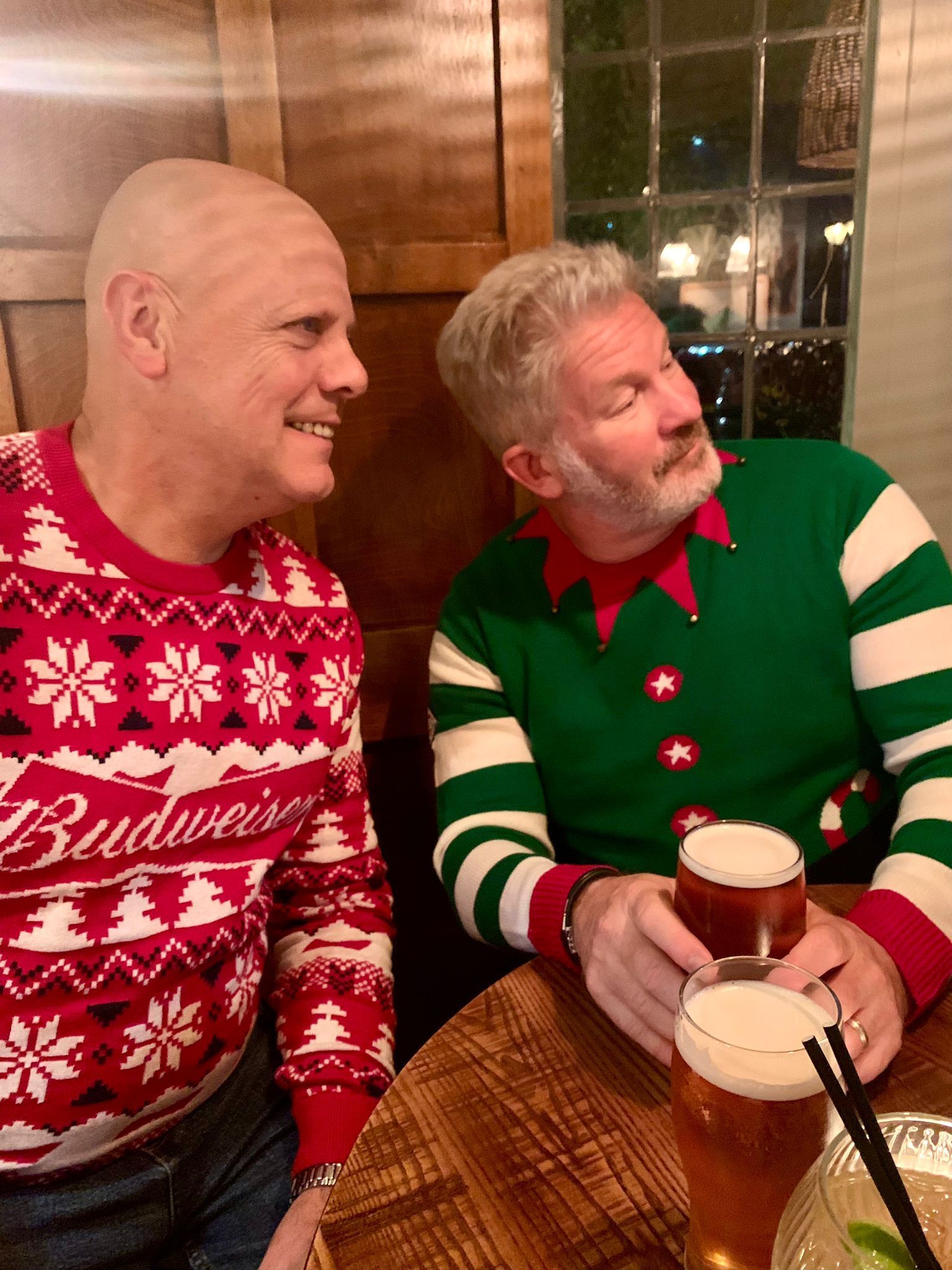 Two people in festive sweaters smiling at a table with drinks in a warmly lit restaurant