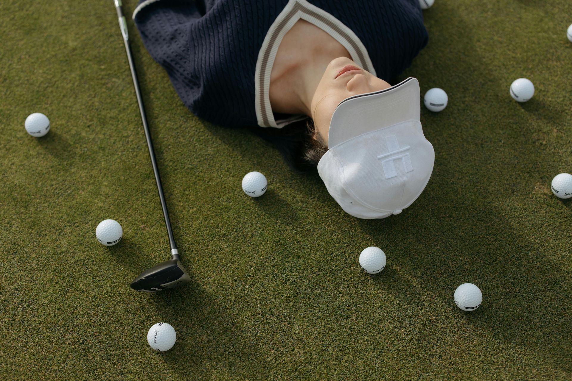 Woman wearing hat, lying on golf green, surrounded by balls, golf club nearby.