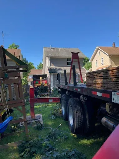 A flatbed truck loaded with lumber sits in a backyard. A playset and houses are in the background.