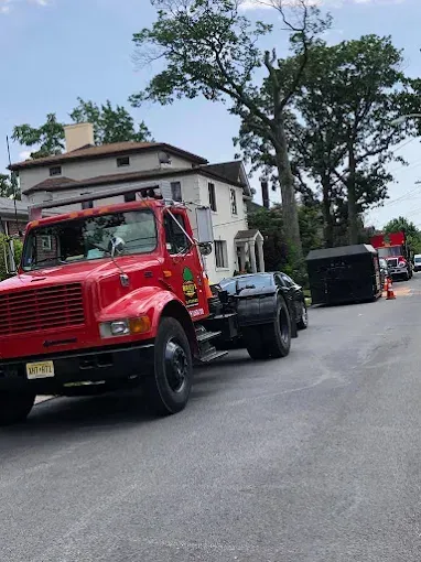 Red truck parked on a street with a black dumpster and a white house in the background.