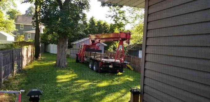 A red crane truck parked in a grassy backyard, likely for construction. Wooden fences and trees surround.