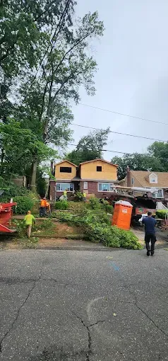 Men trimming fallen tree near a house with an orange porta-potty on a cloudy day.