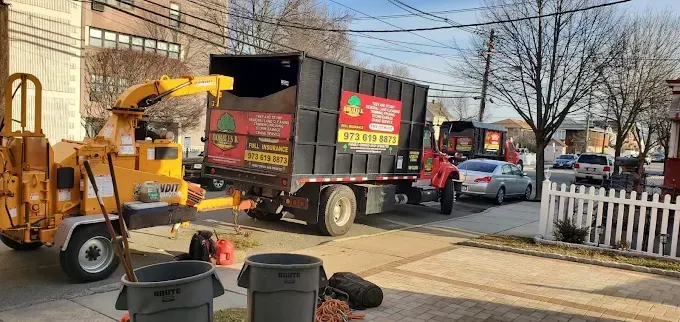 Yellow wood chipper, black dump trucks, and a car parked on the street. Trees and a white picket fence are visible.