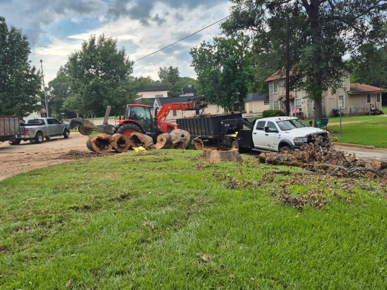 Tractor and dump truck hauling debris beside a white pickup on a muddy residential street after storm cleanup