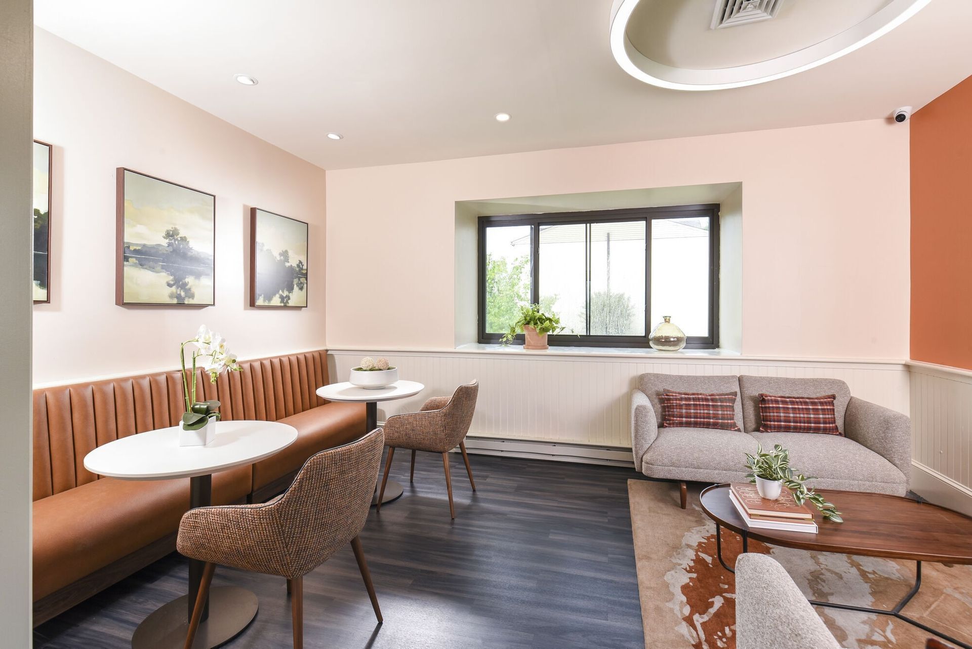 Interior community lounge with pink walls, tan seating, round white tables, and wall art at The Addison, offers townhomes for rent in North Wales, PA.