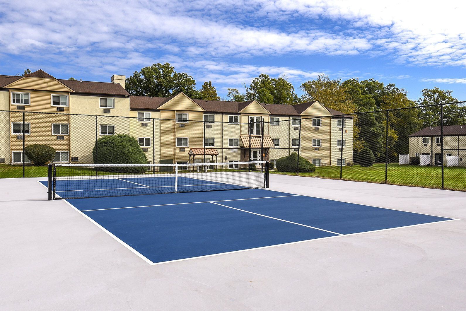Outdoor tennis court with a chain-link fence and apartment buildings beyond at The Addison, offers apartments for rent in North Wales, PA.