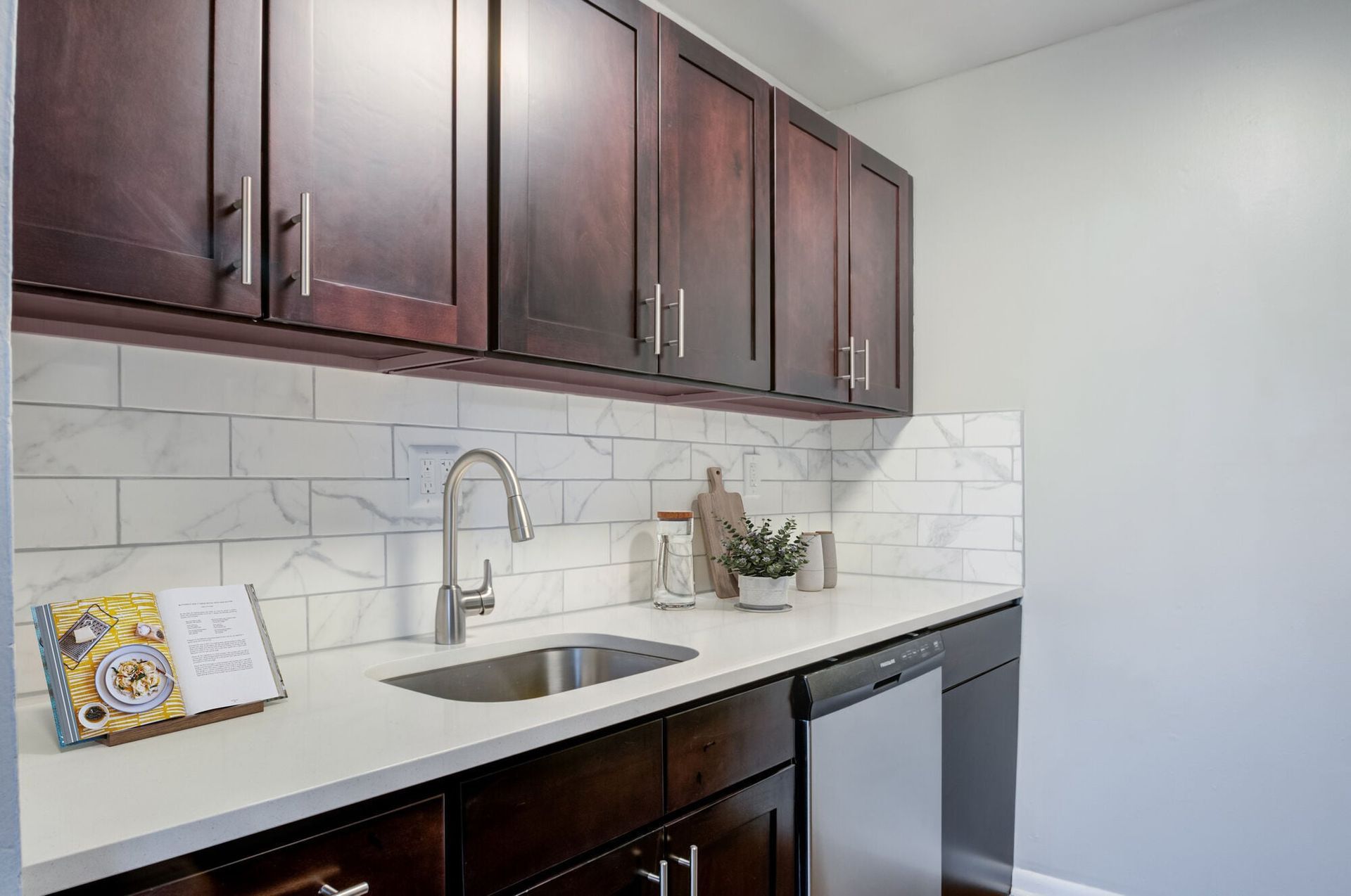Kitchen with dark wood upper cabinets, white countertop, marble backsplash, and stainless steel sink at The Addison, offers apartments in North Wales, PA.