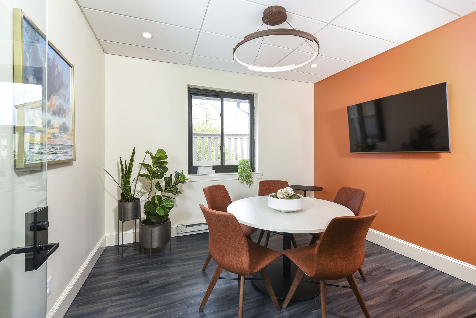 Round white table with rust chairs in a modern community lounge, orange accent wall and wall-mounted TV at The Addison, offers townhomes for rent in North Wales, PA.
