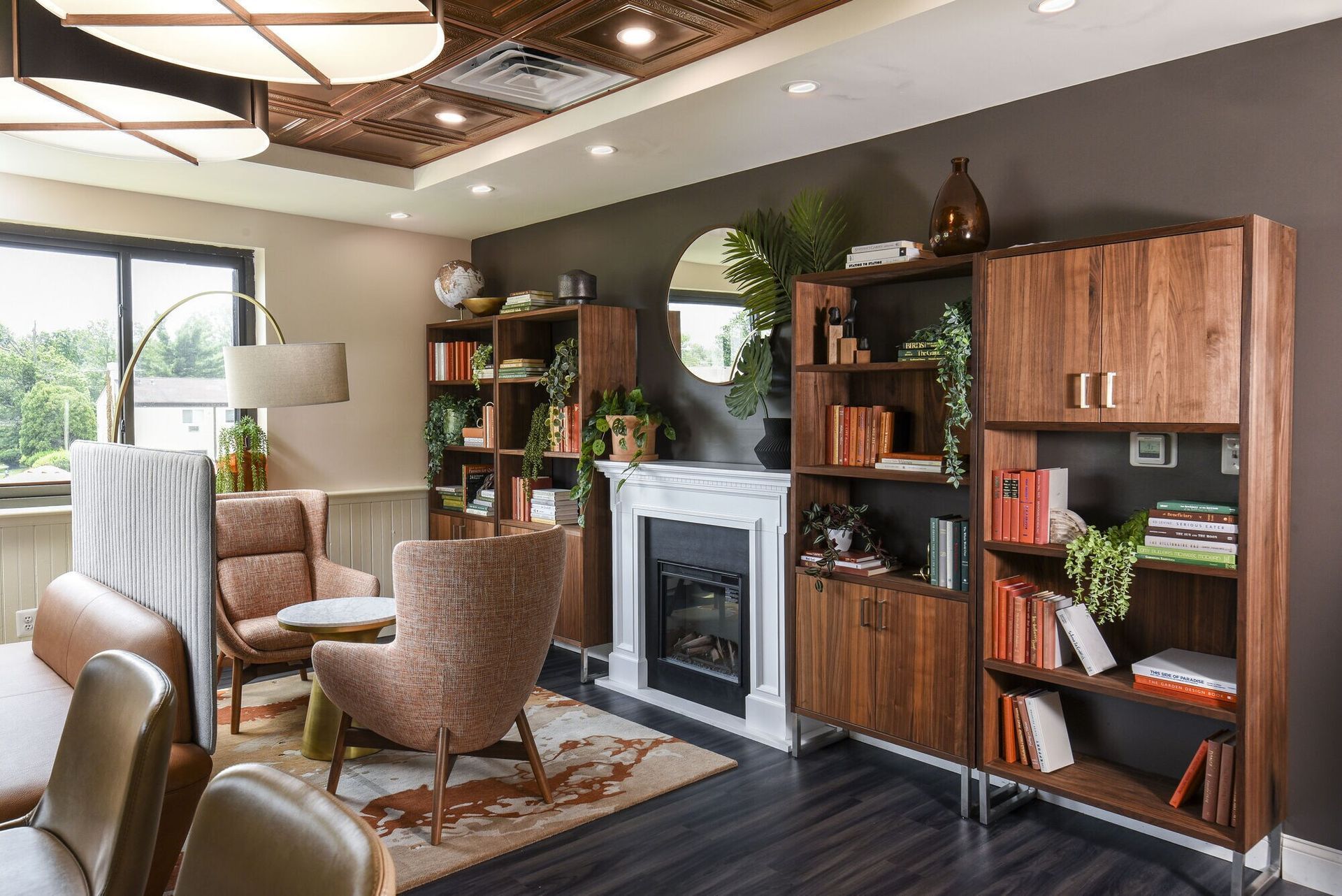 Cozy community lounge with a fireplace, wooden bookshelves, plants, and comfortable chairs at The Addison, offers townhomes for rent in North Wales, PA.