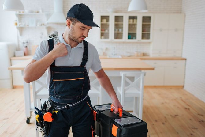 Plumber in a kitchen holding a wrench, looking at a toolbox. Wearing a hat and overalls.