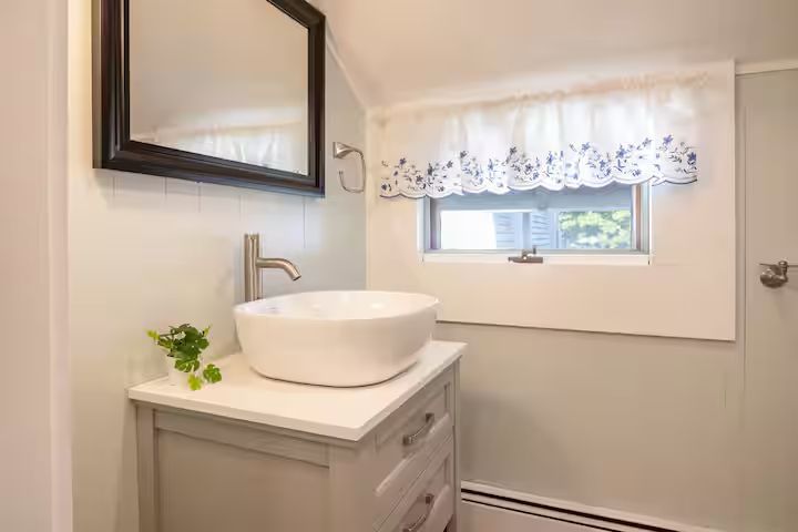 Compact bathroom vanity with round vessel sink, mirror, and small window with lace curtain